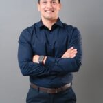 A professional studio portrait of a smiling man with short, dark hair. He is wearing a dark navy blue collared shirt, dark trousers, and a brown leather belt. He is standing with his arms crossed over his chest, and he has a square smart watch visible on his left wrist. The background is a solid, neutral grey