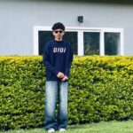 A full-length vertical photo of a young person with dark, curly hair and sunglasses, standing outdoors in front of a hedge and a building. They are wearing a dark navy blue sweater with a light pink graphic that reads "O!O!" and oversized, faded blue denim jeans. The person is standing on a patch of green grass, with a well-maintained, bright green hedge behind them. The background is a plain, light grey wall of a building with a horizontal window